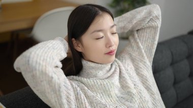 Young chinese woman relaxed with hands on head sitting on sofa at home