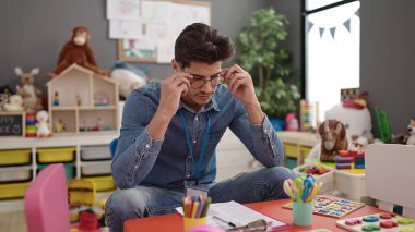 Young hispanic man preschool teacher stressed sitting on table at kindergarten