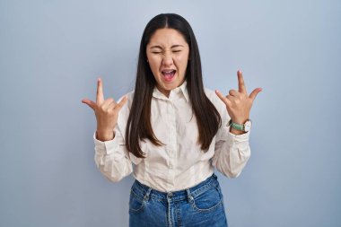 Mavi arka planda dikilmiş çılgın bir ifadeyle bağıran genç Latin kadın elleri havada rock sembolü yapıyor. Müzik yıldızı. ağır müzik konsepti. 