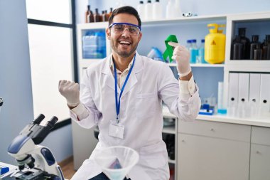 Sakallı genç İspanyol adam elinde yeşil kurdeleyle laboratuvarda çalışıyor gururla bağırıyor, zaferi kutluyor ve kollarını kaldırarak başarıyı kutluyor. 