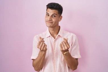 Young hispanic man standing over pink background doing money gesture with hands, asking for salary payment, millionaire business 