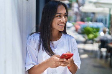 Genç, güzel, İspanyol bir kadın sokakta akıllı telefon kullanarak gülümsüyor.