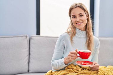 Young blonde woman drinking coffee sitting on sofa at home