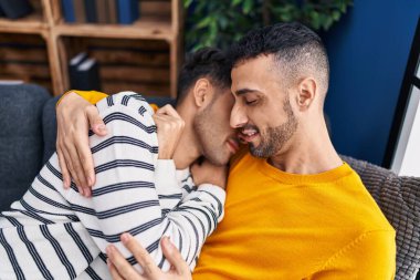 İki erkek, evdeki kanepede oturup birbirlerine sarılıyorlar.