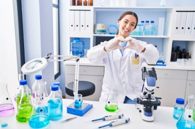 Young brunette woman working at scientist laboratory smiling in love showing heart symbol and shape with hands. romantic concept. 
