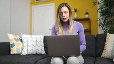 Evdeki kanepede dizüstü bilgisayar kullanan genç sarışın kadın.