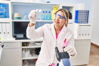 Young hispanic woman wearing scientist uniform looking sample at laboratory
