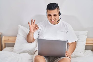 Yatağın üzerinde bilgisayar kullanan İspanyol genç adam parmaklarıyla imza atıyor, gülümseyerek mükemmel bir sembol oluşturuyor. 