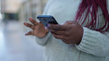 Saçları örgülü Afrikalı bir kadın sokakta akıllı telefon kullanıyor.