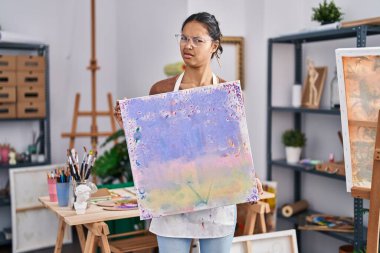 Resim stüdyosunda resim tuvali tutan Brezilyalı genç bir kadın bilgisiz ve kafası karışık bir ifade sergiliyor. şüphe kavramı. 