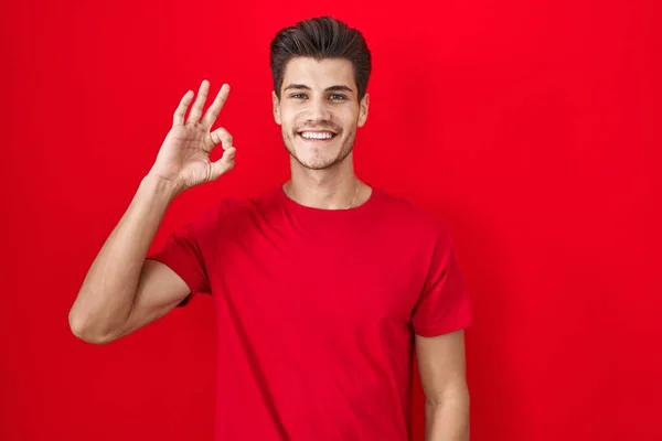 stock image Young hispanic man standing over red background smiling positive doing ok sign with hand and fingers. successful expression. 