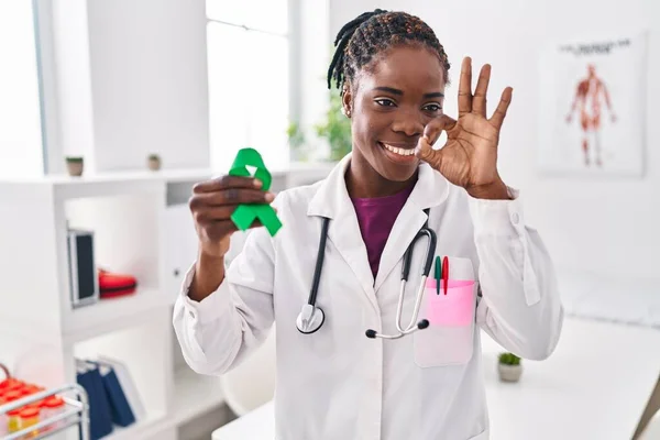 Beautiful black doctor woman holding support green ribbon for mental health awareness doing ok sign with fingers, smiling friendly gesturing excellent symbol