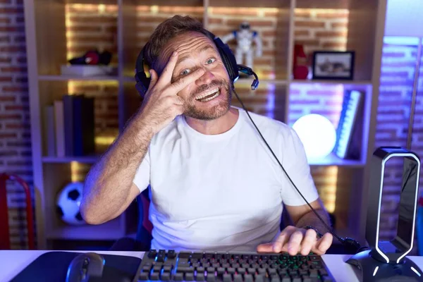 髭を生やした中年の男がビデオゲームをしているヘッドフォンを身に着けている顔の上に指で平和のシンボルを行う 笑顔明るい勝利を示す — ストック写真