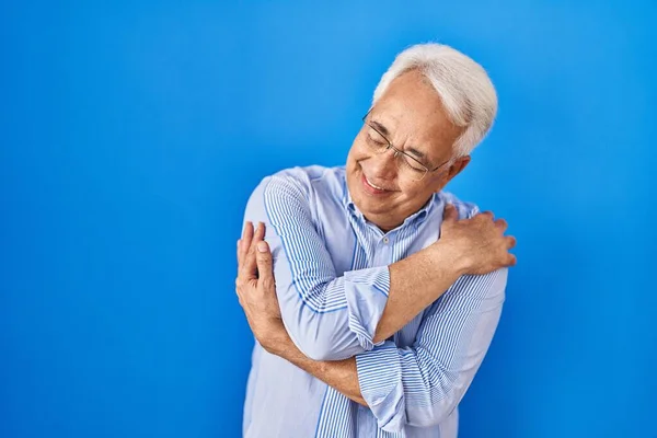 Hispanic senior man wearing glasses hugging oneself happy and positive, smiling confident. self love and self care
