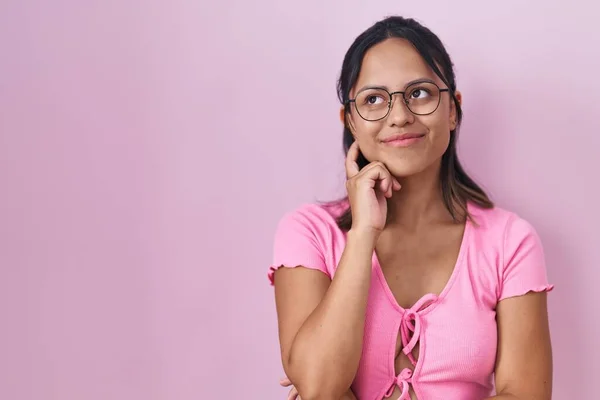 ピンクの背景に立っているヒスパニック系の若い女性の質問については 集中的な表現を考えて手で眼鏡をかけて 思慮深い顔で笑顔 疑わしい概念 — ストック写真