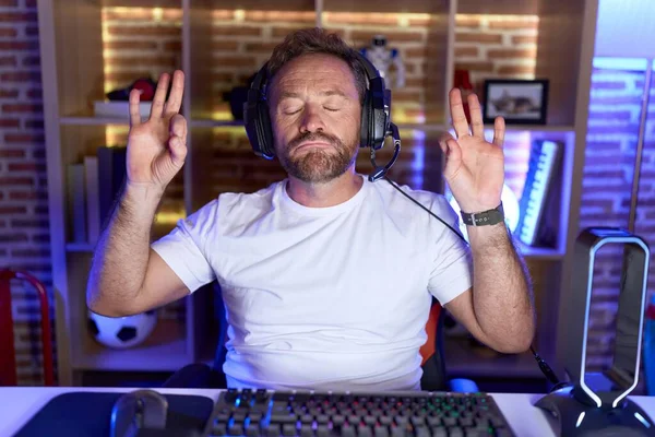髭を生やした中年男性がヘッドフォンでリラックスした笑顔でビデオゲームをして 指で瞑想のジェスチャーをして閉じました ヨガのコンセプト — ストック写真