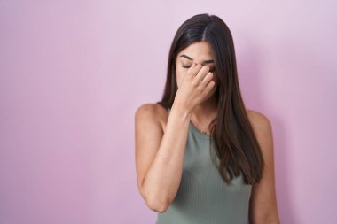 Hispanic woman standing over pink background tired rubbing nose and eyes feeling fatigue and headache. stress and frustration concept. 