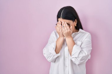 Young brunette woman standing over pink background with sad expression covering face with hands while crying. depression concept. 