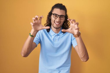 Young hispanic man standing over yellow background smiling funny doing claw gesture as cat, aggressive and sexy expression 