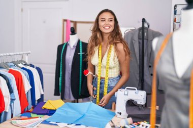 Young beautiful hispanic woman tailor smiling confident cutting cloth at tailor shop