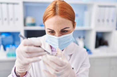 Tıbbi maske takan beyaz kadın bilim adamı laboratuarda cımbızla örnek tutuyor.