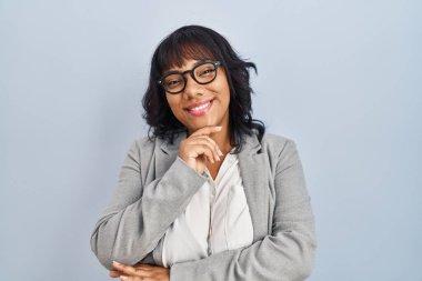 Hispanic woman standing over isolated background looking confident at the camera smiling with crossed arms and hand raised on chin. thinking positive. 