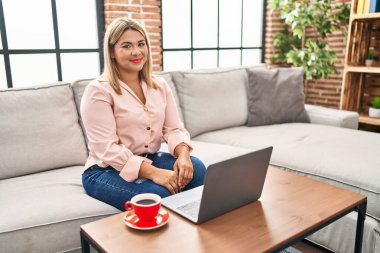Evdeki kanepede dizüstü bilgisayar kullanan genç İspanyol kadın yüzünde ciddi bir ifadeyle rahatlamış olarak oturuyor. Basit ve doğal bir şekilde kameraya bakıyorsun.. 