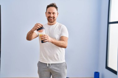 Spor merkezinde kendinden emin gülümseyen beyaz bir adam protein içeceği sallıyor.