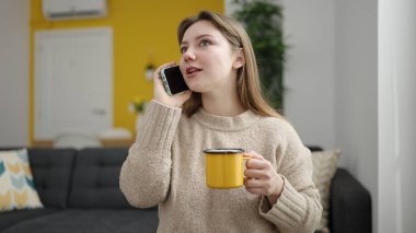 Young blonde woman talking on smartphone drinking coffee at home