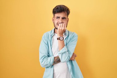 Sarı arka planda dikilen İspanyol dövmeli genç adam elleri ağız ısıran tırnaklarıyla stresli ve gergin görünüyor. anksiyete. 