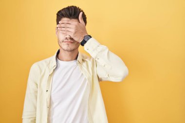 Sarı arka planda dikilen İspanyol genç adam gözlerini eliyle kapatıyor, ciddi ve üzgün görünüyor. Görme, saklama ve reddetme konsepti 