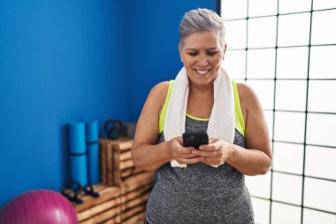 Spor kıyafetleri giyen orta yaşlı bir kadın spor merkezinde akıllı telefon kullanıyor.