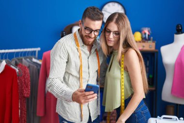 Man and woman tailors smiling confident using smartphone at clothing factory