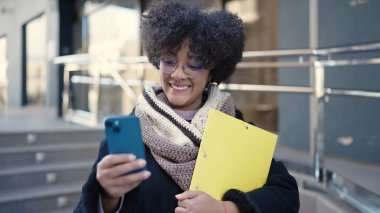 Genç Afro-Amerikalı kadın gülümsüyor. Elinde klasör ile caddede akıllı telefon kullanıyor.