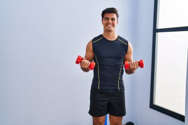 Young hispanic man smiling confident using dumbbells training at sport center