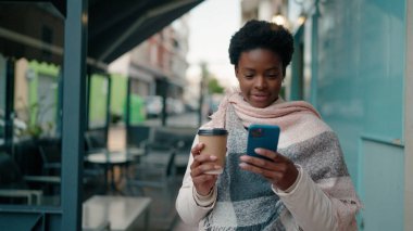 Genç Afrikalı Amerikalı kadın akıllı telefon kullanıyor. Sokakta kahve içiyor.