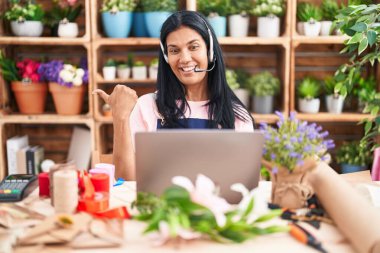 Çiçekçide çalışan olgun İspanyol bir kadın video çekiyor. Başparmağıyla işaret ediyor. Ağzı açık gülümsüyor. 