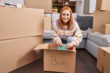 Beyaz bir kadın gülümsüyor. Yeni evindeki karton kutuları açıyor.