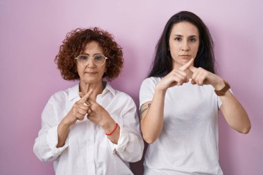 Hispanic mother and daughter wearing casual white t shirt over pink background rejection expression crossing fingers doing negative sign 