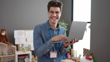 Young hispanic man preschool teacher using laptop standing at kindergarten
