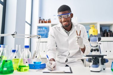 Young african american man wearing scientist uniform writing on clipboard holding test tubes at laboratory