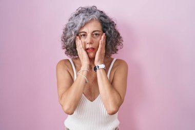 Pembe arka planda duran, gri saçlı orta yaşlı bir kadın. Yüzü, depresyonu ve üzüntüyü örten yorgun elleri var. 
