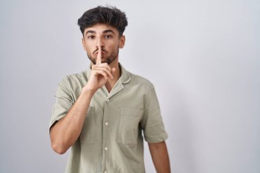 Beyaz arka planda duran sakallı bir Arap, parmakları dudaklarında sessiz olmak istiyor. sessizlik ve gizli konsept. 