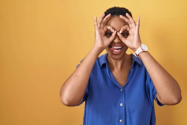 非洲裔美国妇女站在黄色的背景上做着像双筒望远镜一样的手势 伸出舌头 眼睛透过手指看着 疯狂的表达 — 图库照片