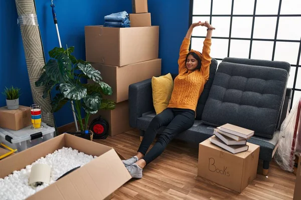 Young latin woman stretching arms relaxed on sofa at new home