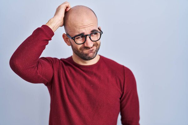 Young bald man with beard standing over white background wearing glasses confuse and wondering about question. uncertain with doubt, thinking with hand on head. pensive concept. 