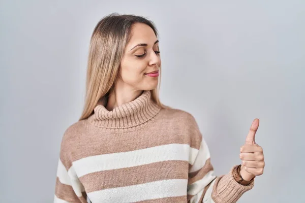 Young blonde woman wearing turtleneck sweater over isolated background looking proud, smiling doing thumbs up gesture to the side