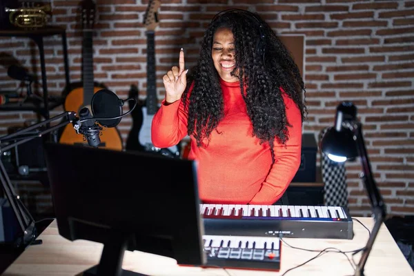 stock image Plus size hispanic woman playing piano at music studio showing and pointing up with finger number one while smiling confident and happy. 