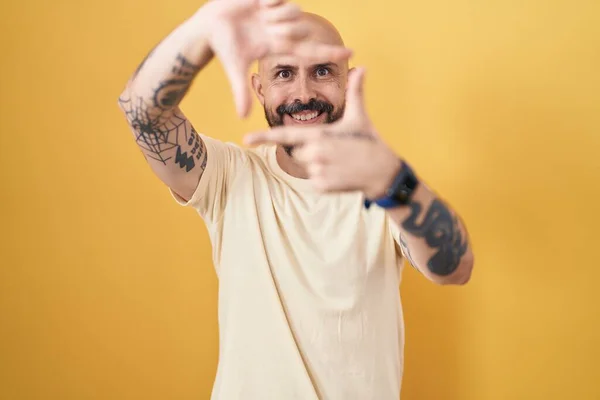 Hispanic man with tattoos standing over yellow background smiling making frame with hands and fingers with happy face. creativity and photography concept.
