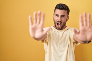 Yakışıklı İspanyol adam sarı arka planda duruyor. Avuçlarıyla jesti durduruyor. Öfkeli ve hayal kırıklığı dolu bir ifade. 